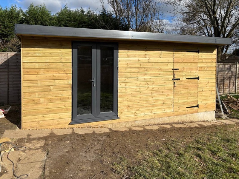  Exterior side view of a completed garden room with natural wood horizontal siding, black-framed double doors, and a matching side storage door. 