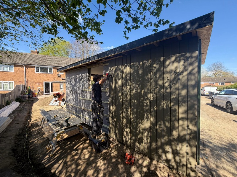  A carpenter installing vertical composite cladding onto a garden room structure, with the moisture barrier and timber battening still visible on the unclad section. 