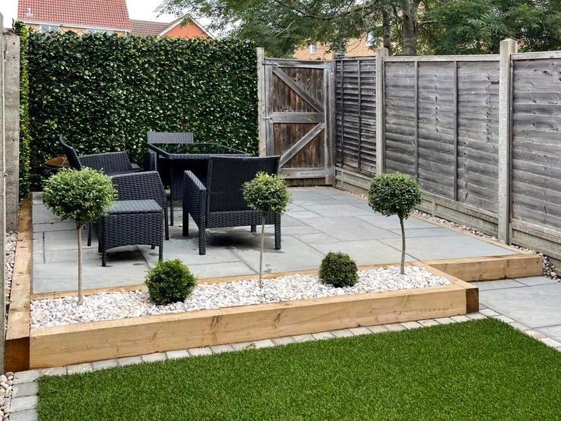 A beautiful patio area at the end of a small garden with a black cane style table and three chairs and surrounded by a fence and high hedge